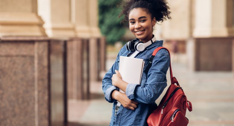 Cheerful Black Student Girl Hugging Books Posing With Backpack Near College Building Outdoor, Smiling To Camera. Modern Education, College Tuition And Grants, Studentship And Study Abroad Concept