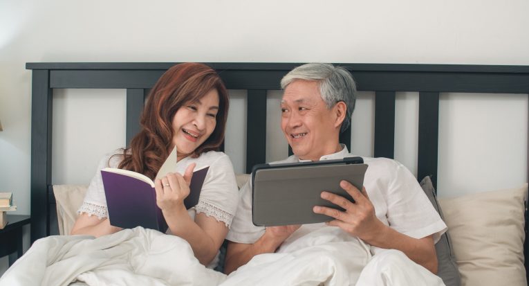 Asian senior couple using tablet at home. Asian Senior Chinese grandparents, husband watch movie and wife read book after wake up, lying on bed in bedroom at home in the morning concept.