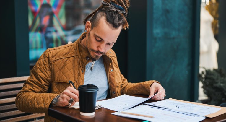 Handsome guy with dreadlocks is relaxing with coffee outdoors while reading and making notes in sketchbook