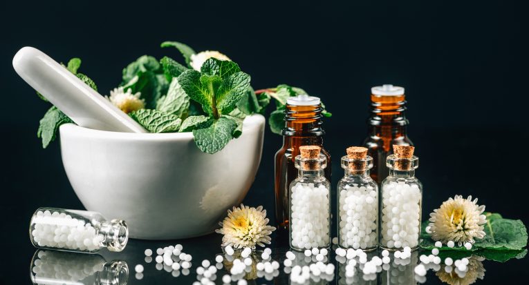 Homeopathy Treatment - Homeopathic globules scattered out of glass bottle, mortar and pestle full of fresh medicinal plants, black background