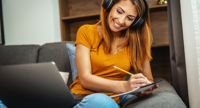 Cute young female student sitting on the sofa, using her laptop and headphones to watching lesson online and learning from home. Young woman taking notes while following teacher doing lesson on video call.