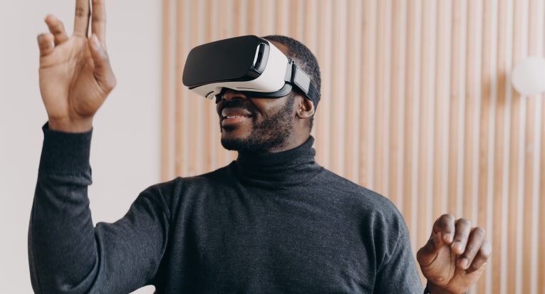 Young smiling african american businessman in VR glasses sitting in office, testing virtual reality goggles for business, impressed man using 3D technology, touching objects in air with hands