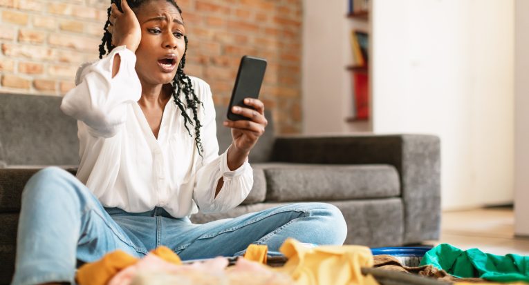 Terrified african american woman staring at smartphone screen in terror, feeling shocked about cancelled flight, sitting near open suitcase at home, free space