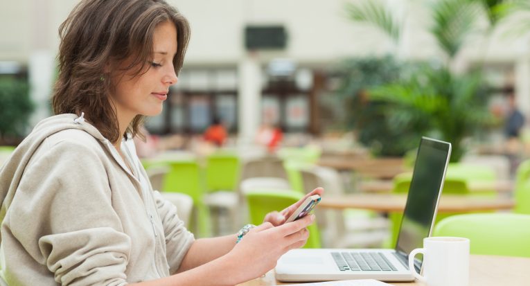 Side view portrait of a female student using cellphone and laptop at cafeteria table