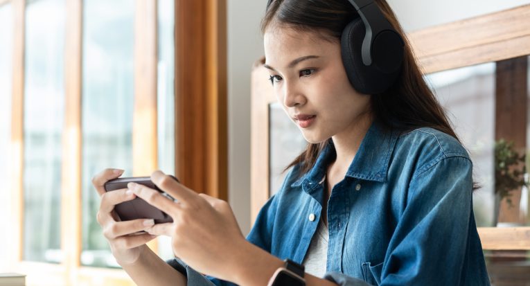 Young woman student in headphone is using smartphone to playing games and listening to the music after reading hard textbook to preparing for examination in college.