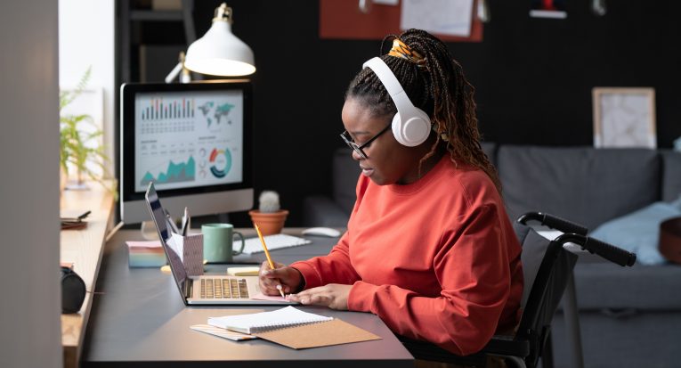 African girl in wireless headphones sitting at the table in front of the laptop and making notes, she studying at home