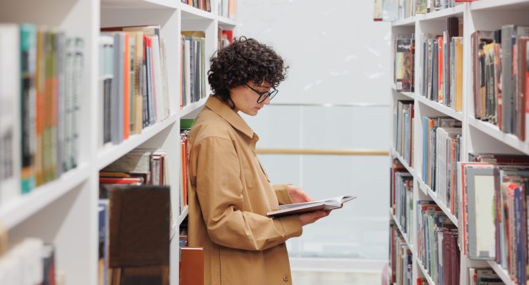young woman with curly hair, library among the shelves books. female student conducts scientific work or writes diploma. happy confident woman with books in hands. work and education
